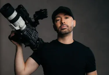 A person wearing a black t-shirt holds a professional camera rig, with a plant in the background against a gray backdrop