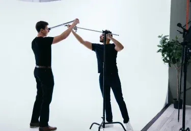 Two men in black shirts collaborate to adjust equipment on a stand in a bright, minimalist studio setting
